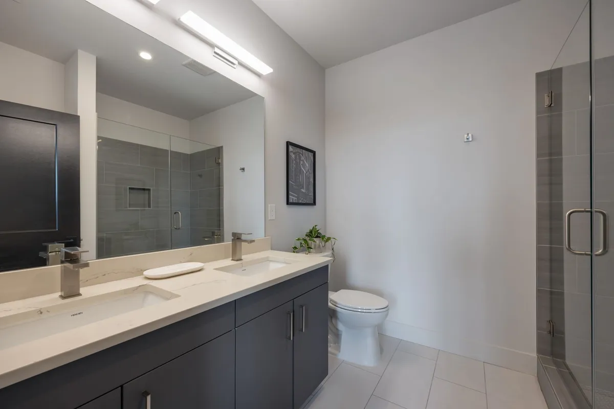 A spa-inspired en-suite bathroom with double sink vanity, a large mirror, a spacious shower area with glass enclosure, and a toilet. The clean lines and bright ambiance evoke a sense of calm and sophistication.