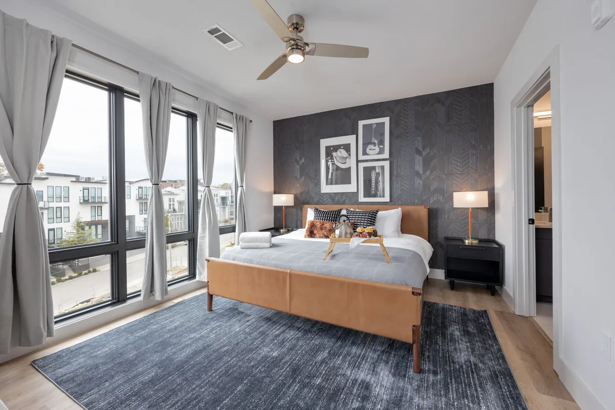A serene bedroom featuring a plush bed, large sun-filled windows, tasteful artwork on the wall, and nightstands topped with soft-glow lamps. The space is inviting and tranquil, highlighting both comfort and style.