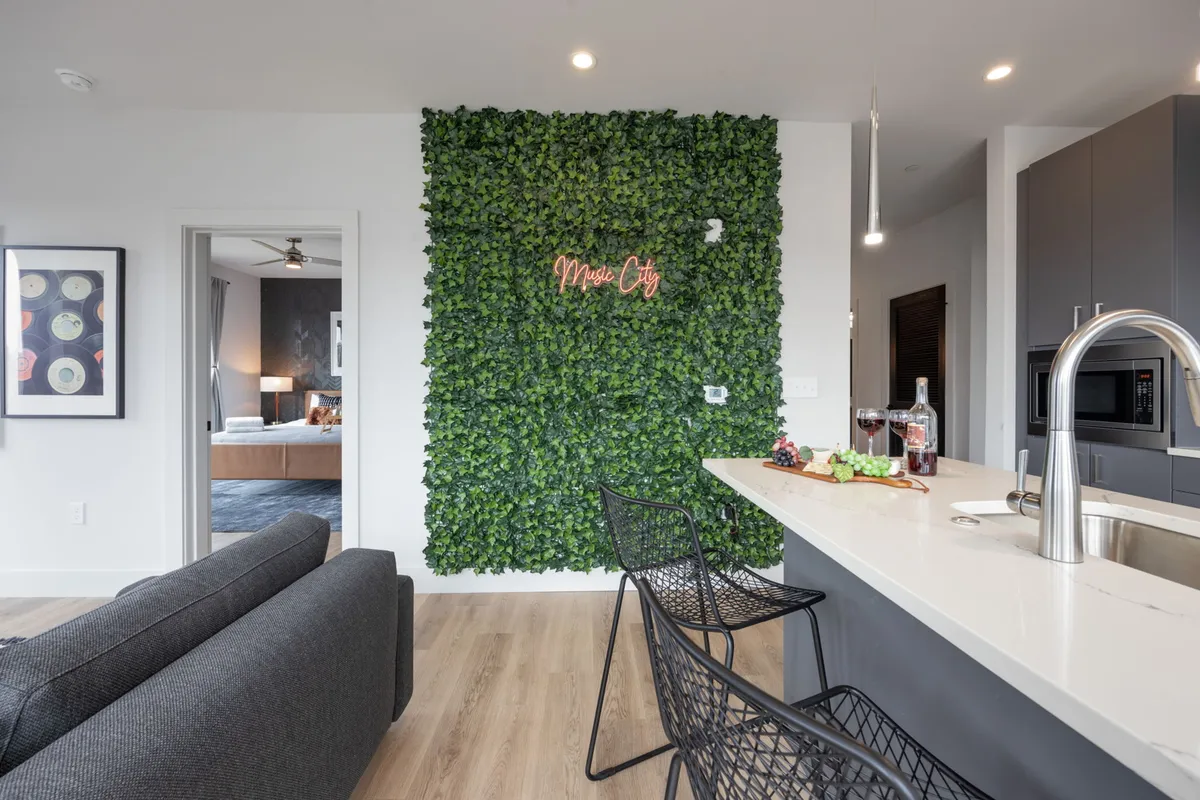 A lively and contemporary indoor common area featuring a wall of greenery, a neon sign, a stylish bar, and a comfortable sitting area. The space is designed for both social gatherings and relaxation, exuding a welcoming ambiance.