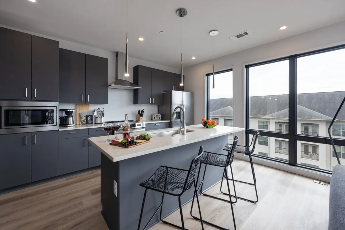 The photo showcases a sleek, contemporary kitchen with an island countertop, bar stools, large windows, and modern appliances. The space is naturally lit and set up for cooking, entertaining, and casual dining.