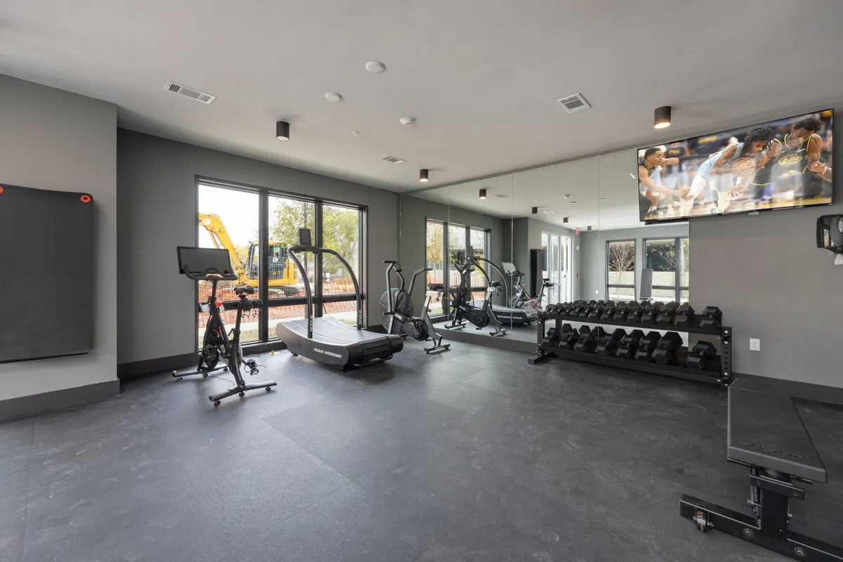 A bright, modern fitness center equipped with treadmills, elliptical machines, a dumbbell rack, and large windows that let in natural light. A TV screen is mounted for added convenience, and the overall atmosphere is energetic and motivating.