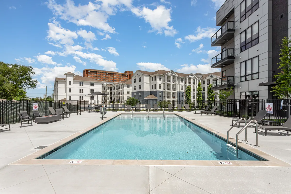 A sunlit swimming pool surrounded by contemporary apartment buildings, with a spacious poolside seating area and lush green landscaping. The setting is lively yet relaxing, suggesting opportunities for both social gatherings and peaceful sunset swims.