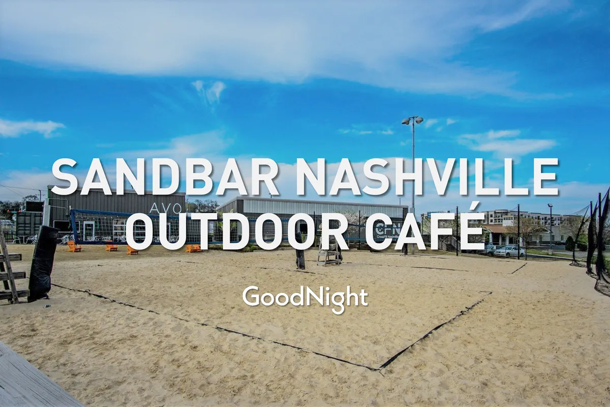 A lively outdoor café scene featuring a sand volleyball court, plenty of seating, and a sky backdrop. The area is bustling with friends socializing, playing, and relaxing, evoking a vibrant lakeside or park-like atmosphere.