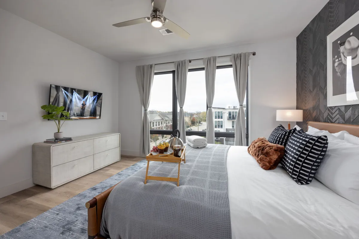 A serene bedroom featuring a plush king bed adorned with decorative pillows, a tray with breakfast items on the bed, a smart TV mounted on the wall, and large windows that allow natural light to stream in. The décor blends modern comfort with inviting textures, creating a restful atmosphere.