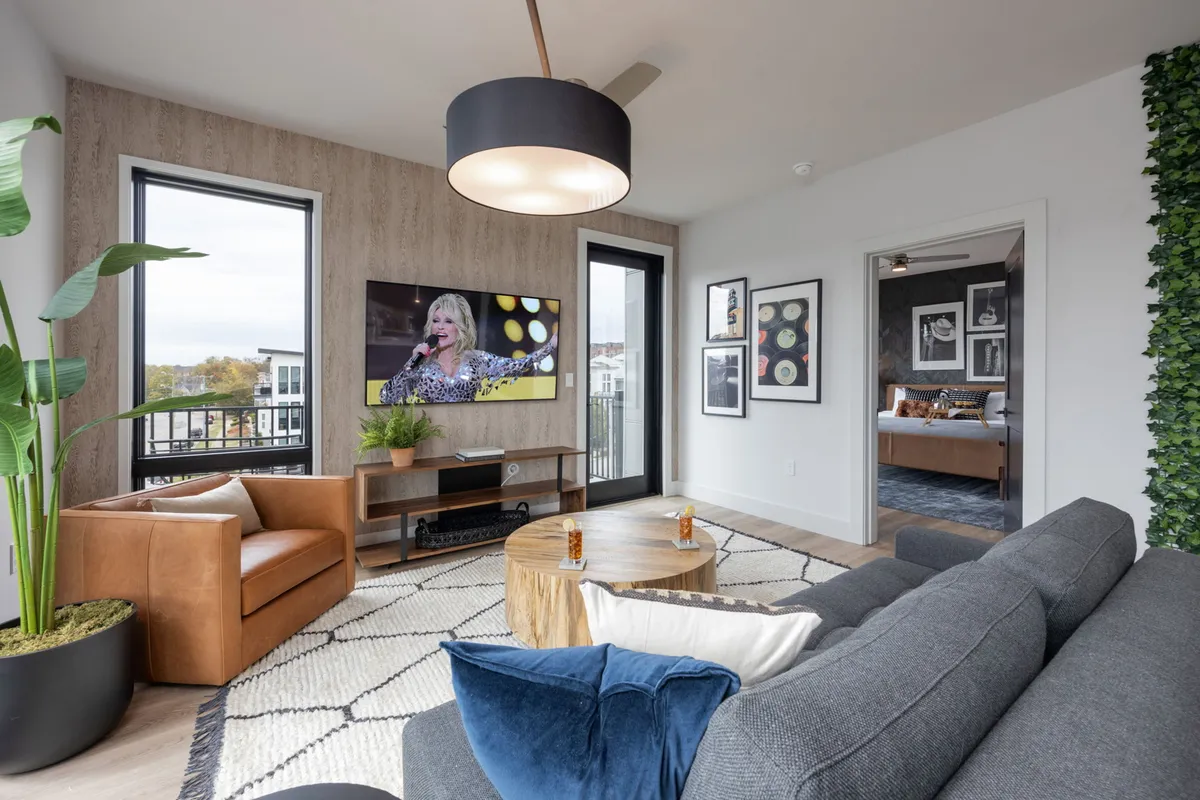 The image shows a spacious and inviting living room with a large, plush sectional sofa arranged around a sleek wooden coffee table. A television is mounted on the wall, currently displaying a lively performance, adding to the entertainment-friendly vibe. The room is accented with a vibrant green plant and thoughtfully selected framed artwork, creating a stylish yet cozy atmosphere perfect for gathering, relaxing, or entertaining.