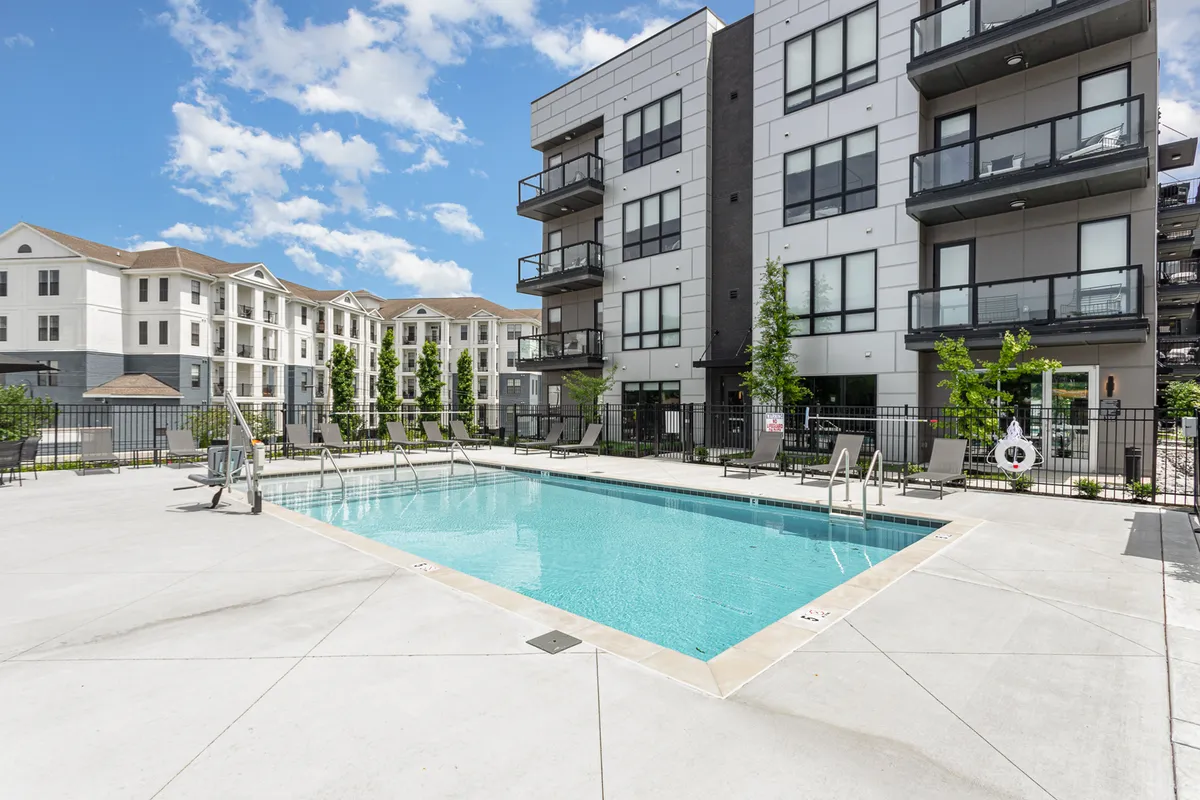 A sunlit swimming pool surrounded by landscaped greenery and the balconies of a modern building, inviting both relaxation and social activity. The pool area is peaceful, with hints of community interaction.